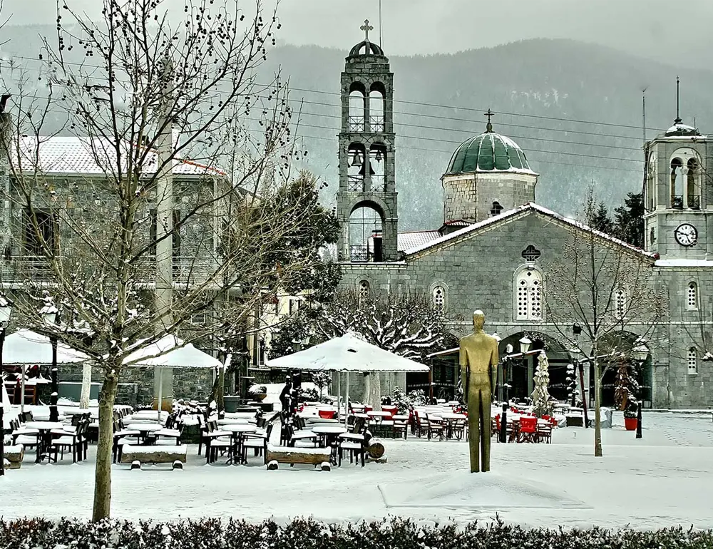 Χιονισμένη πλατεία της Βυτίνας με την εκκλησία του Αγίου Τρύφωνα και τα πέτρινα καμπαναριά.